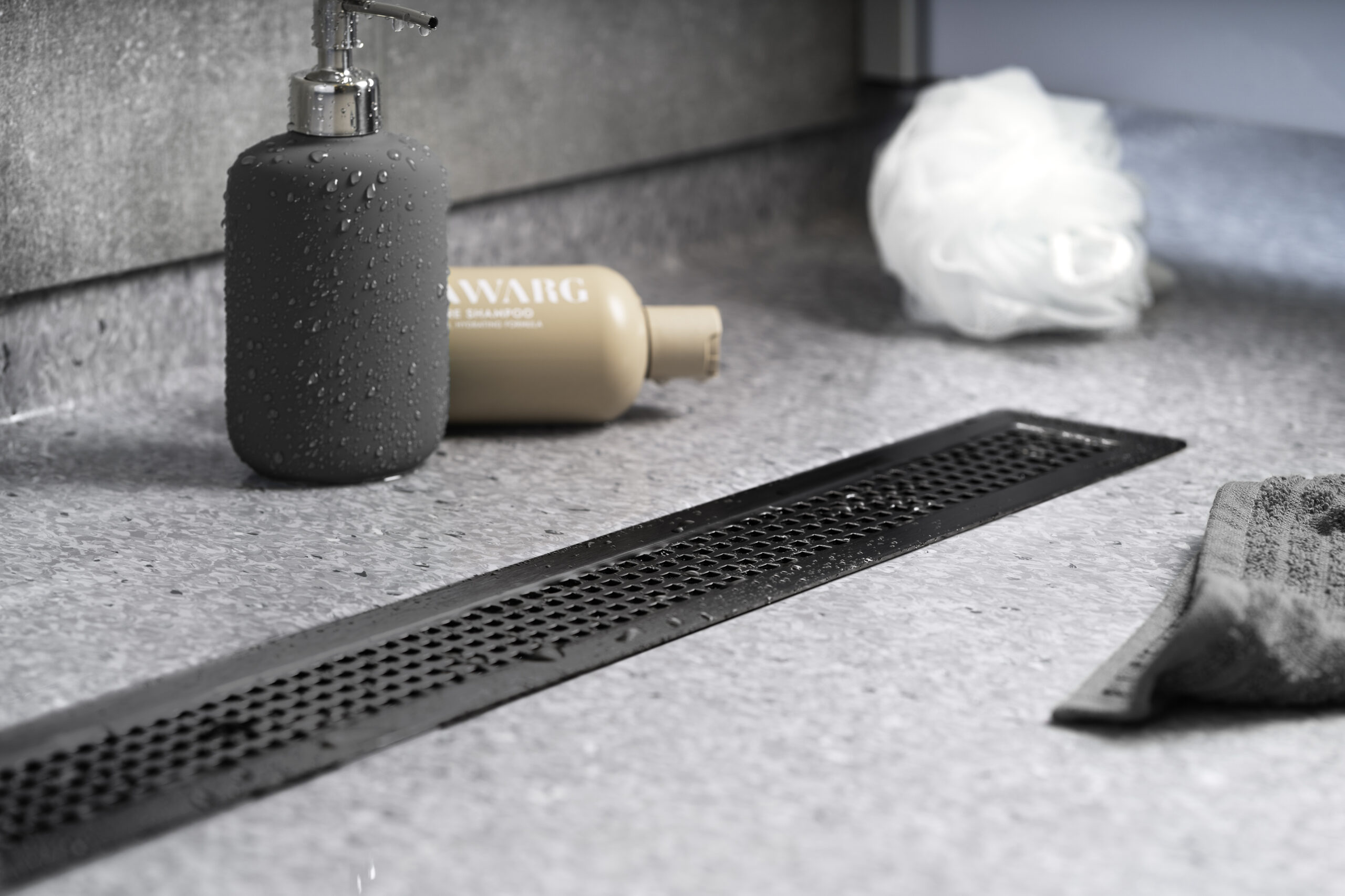 Et nærbilde av matt svart Purus Line Vinyl Plus designsluk, vakkert integrert i et grått gulvbelegg, viser en sømløs og elegant løsning for moderne baderom med vinylgulv.