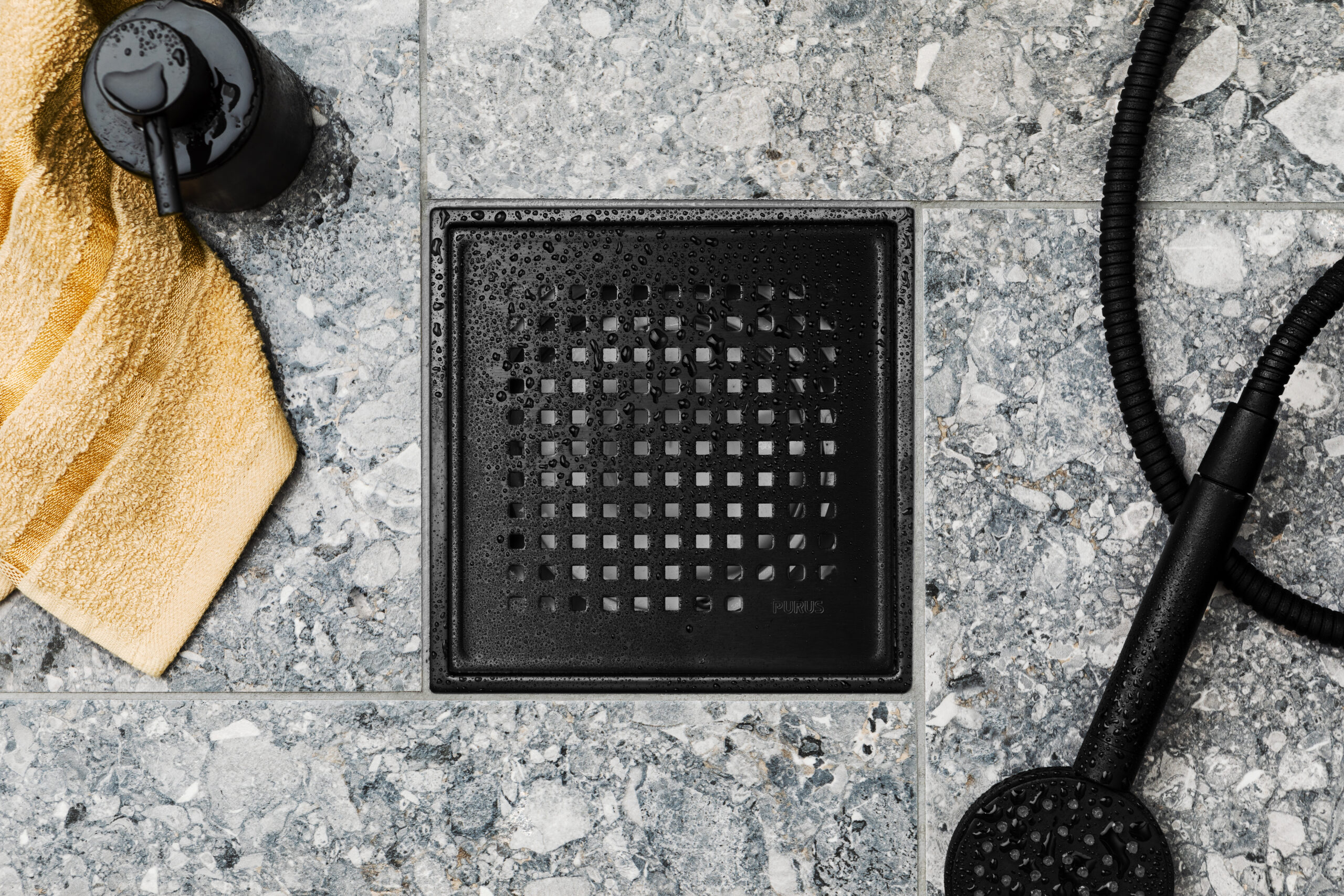 Et nærbilde av Purus Square matt svart slukrist i Chess-mønster, elegant plassert i et flislagt gulv med grå Ceppo/terrazzo-fliser, viser et moderne og stilfullt designvalg for ethvert baderom.