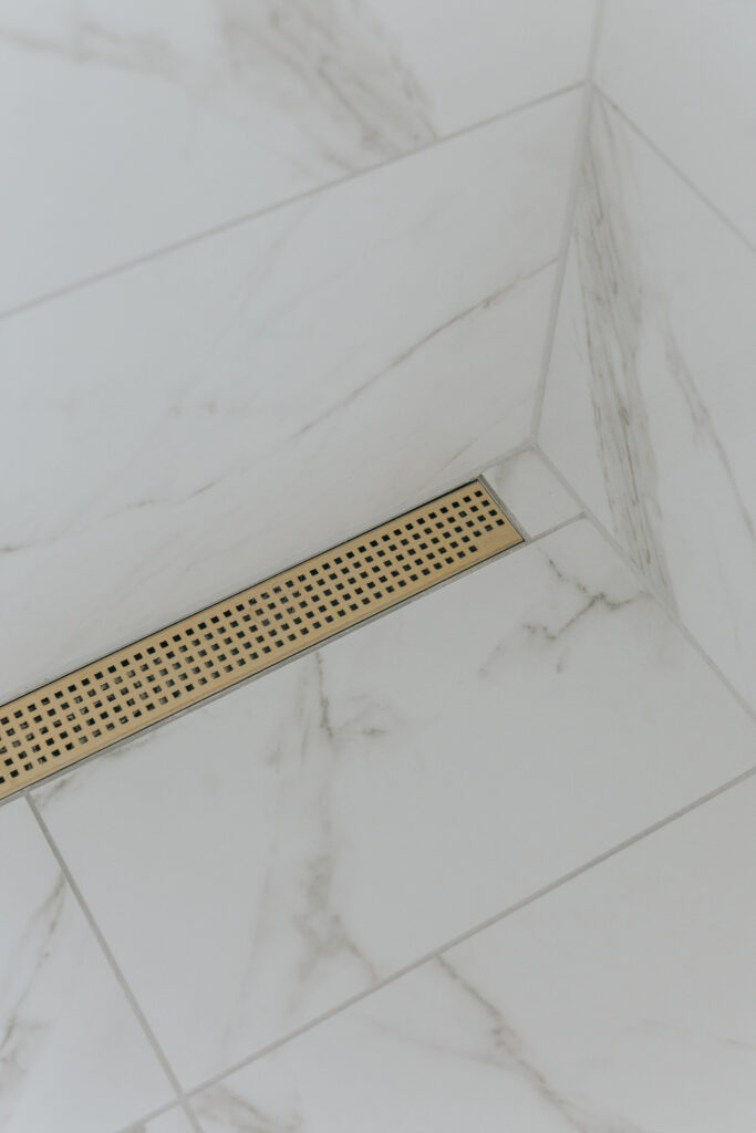 Nærbilde av slukrennen Purus Line Chess, designsluk, med blank gulloverflate. Store marmorfliser er lagt i dusjsonen - på både vegg og gulv, og gir en behagelig spa-følelse på badet.